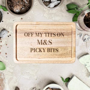 Puede incluir: Una tabla de cortar rectangular de madera con un borde tallado. El texto "OFF MY TITS ON M&S PICKY BITS" está grabado en la tabla. La tabla está rodeada de cuencos pequeños, queso y una copa de vino tinto.