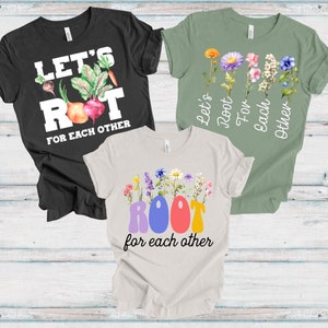 May include: Three t-shirts with the slogan "Let's Root For Each Other." One black shirt features vegetables, another green shirt has flowers, and the third is beige with colourful letters and flowers. All shirts are short-sleeved.