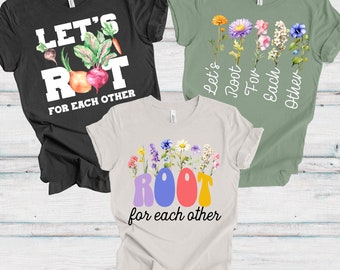 Può includere: Tre magliette con la frase "Let's Root For Each Other". Una maglietta nera con verdure, un'altra verde con fiori e la terza beige con lettere e fiori colorati. Tutte le magliette sono a maniche corte.