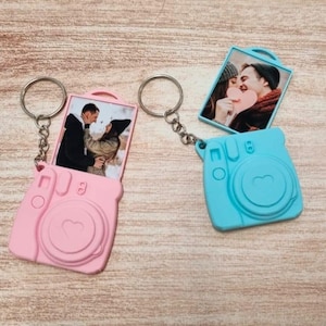 Puede incluir: Dos llaveros en forma de cámara rosa y azul con anillas. Cada llavero tiene un inserto para fotos con una pareja en una pose romántica.