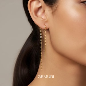 May include: A pair of gold-colored earrings. One earring features a delicate chain, while the other has a straight bar pendant. The earrings are shown on a person's ear. The word "GEMURI" is printed at the bottom.