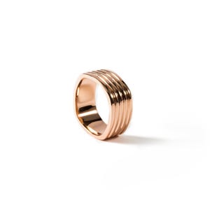 Könnte beinhalten: Ein roségoldener Ring mit geripptem Design. Der Ring hat ein breites Band mit mehreren parallelen Rillen, wodurch eine strukturierte Oberfläche entsteht. Der Ring ist vor einem weißen Hintergrund platziert, der seinen metallischen Glanz und seine komplizierten Details hervorhebt.