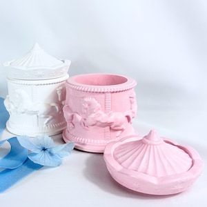 Può includere: Contenitori decorativi a tema giostra, bianchi e rosa. Il contenitore bianco ha un coperchio, e quello rosa è aperto. Entrambi presentano cavalli della giostra in rilievo. Un nastro blu e un fiore sono in primo piano.