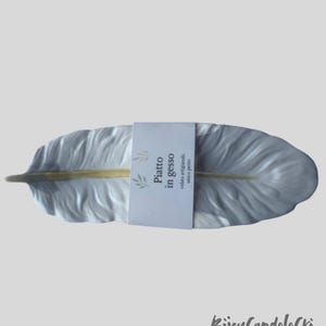 Op de afbeelding: Een witte, veervormige decoratieve schaal met een gouden accent in het midden. De schaal heeft een textuur en een papieren label met Italiaanse tekst. De schaal is een uniek stuk.