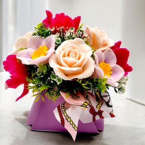 May include: A bouquet of pink, white, and red flowers arranged in a purple box with a patterned ribbon. The flowers are made of soap.