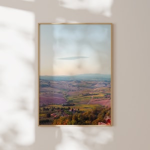 Può includere: Una stampa incorniciata raffigurante un paesaggio pittoresco di dolci colline e valli sotto un cielo morbido e nuvoloso. L'opera presenta una tavolozza di verdi, marroni e viola, con una cornice dorata.