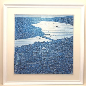 May include: A framed blue and white artwork depicting a detailed cityscape with a bridge and water. The artwork is in a white frame and has a cream-colored mat. The image is a detailed illustration of a city.