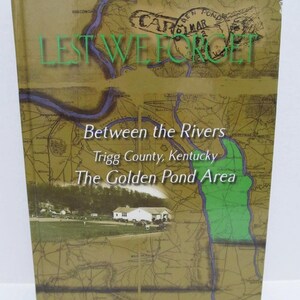May include: A book cover with the title "Lest We Forget" in green letters. The book's title is over a map with the text "Between the Rivers, Trigg County, Kentucky, The Golden Pond Area". The cover has a vintage aesthetic.