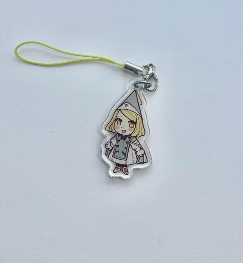 May include: A clear acrylic charm with a cartoon illustration of a character in a green and white outfit and hat. The charm has a yellow lanyard attached with a silver clasp.