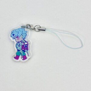 May include: A clear acrylic charm featuring a cartoon character with blue hair, sunglasses, and a backpack. The character is wearing a green and purple outfit and pink boots. The charm has a silver clasp and a light blue lanyard.