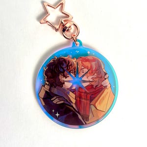 Peut inclure: Un porte-clés rond et irisé avec une illustration de deux personnages s'embrassant, avec une étoile bleue entre eux. Le porte-clés a un fermoir en forme d'étoile de couleur or rose. Les personnages ont les cheveux bruns et roux et portent des manteaux et des écharpes.