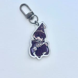 May include: A clear acrylic keyring featuring a cartoon wizard in a purple robe and hat. The wizard has dark hair and is holding a small object. The keyring has a silver-coloured clasp.