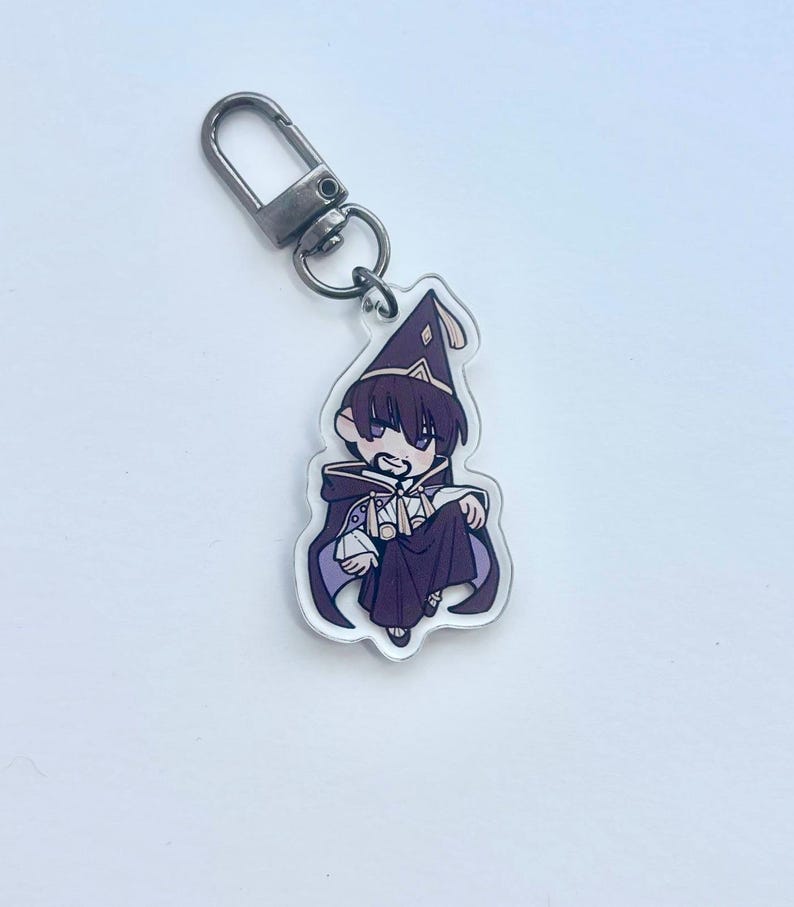 May include: A clear acrylic keyring featuring a cartoon wizard in a purple robe and hat. The wizard has dark hair and is holding a small object. The keyring has a silver-coloured clasp.