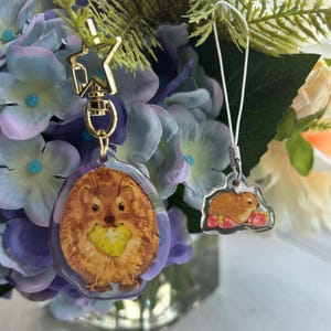 Puede incluir: Dos llaveros de acrílico transparente con herrajes dorados. Un llavero presenta un erizo marrón con una fruta amarilla, el otro un pequeño animal marrón sobre fresas rojas. Ambos sobre un fondo de flores moradas y blancas.