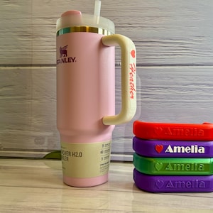 May include: A pink Stanley H2.0 FlowState 40 oz tumbler with a white handle and straw. The handle is personalized with the name "Heather" and a heart. Stacked next to the tumbler are four colored handle wraps with the name "Amelia".