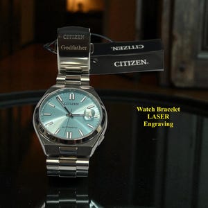 Puede incluir: Un reloj Citizen con esfera azul claro y correa plateada. El reloj tiene la palabra "Citizen" en la esfera y en la parte trasera. El reloj está etiquetado como "Automatic". El reloj se muestra con una etiqueta que dice "Citizen" y "Godfather". También es visible el texto "Watch Bracelet LASER Engraving".