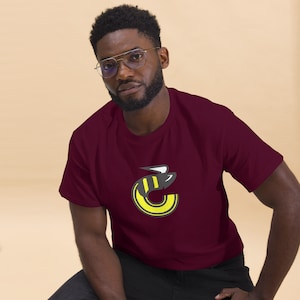 Puede incluir: Una camiseta color granate con un gráfico de abeja amarillo y negro en la parte delantera. La abeja está estilizada para parecerse a la letra "C".