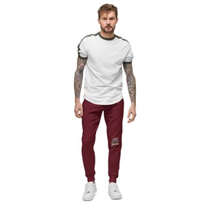 May include: A man wearing a white t-shirt with green trim on the sleeves and burgundy sweatpants with a graphic print on the left leg. He is wearing white sneakers.