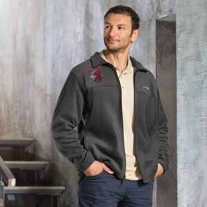 May include: A dark gray fleece jacket with a zipper closure. The jacket has a red and gray embroidered logo on the left chest that says "Fury".