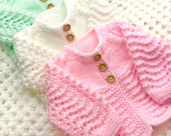 May include: Three knitted baby sweaters in pink, white, and light green. The sweaters have a delicate lace pattern and are fastened with wooden buttons.