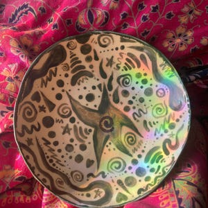 May include: A ceramic bowl with a white background and brown and green abstract designs. The bowl has a holographic finish that creates a rainbow effect.