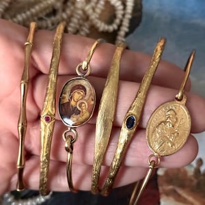 May include: A collection of gold-toned bracelets, each with unique designs. Some feature textured surfaces, while others have decorative elements like gemstones and religious iconography. The bracelets vary in style, offering a diverse range of options.