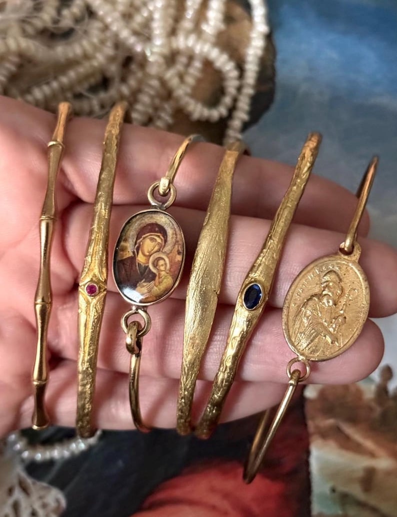 May include: A collection of gold-toned bracelets, each with unique designs. Some feature textured surfaces, while others have decorative elements like gemstones and religious iconography. The bracelets vary in style, offering a diverse range of options.