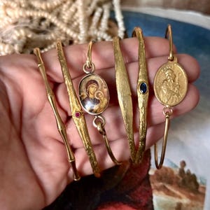 May include: A collection of gold-toned bracelets, each with unique designs. One features a bamboo-like texture, others have oval pendants with religious imagery, and one has a blue gemstone. The bracelets are displayed on a hand.