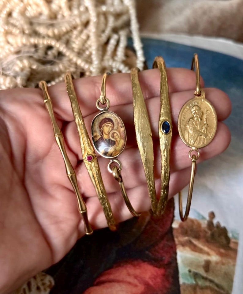 May include: A collection of gold-toned bracelets, each with unique designs. One features a bamboo-like texture, others have oval pendants with religious imagery, and one has a blue gemstone. The bracelets are displayed on a hand.