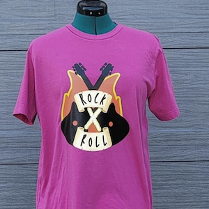 Pode incluir: Uma t-shirt magenta com um design gráfico com guitarras cruzadas, chamas e as palavras "Rock X Roll" numa faixa. O design é em tons de preto, castanho, amarelo e branco. A t-shirt é exibida num manequim.