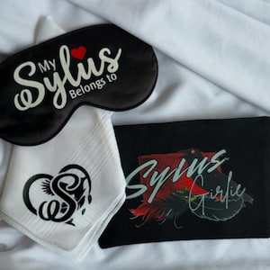Puede incluir: Una máscara para los ojos negra con el texto "My Sylus Belongs to" y un corazón rojo. Un pañuelo blanco con un diseño negro y una bolsa negra con el texto "Sylus Girlie" y un gráfico de pájaro.