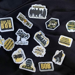 May include: A selection of stickers with diverse designs and text, such as "Stop Val and save BOB", "We are the THUNDERBOLTS", and "TEAM!". The stickers are predominantly black and yellow in colour.