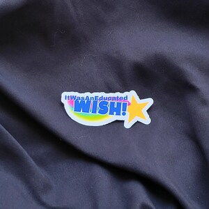 以下が含まれることがあります： 虹色の弧と黄色の星が付いた白いステッカー。ステッカーには「It Was An Educated WISH!」と書かれています。