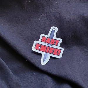 以下が含まれることがあります： 黒い布の背景に、黒いナイフと赤い文字で「BABY KNIFE！」と書かれた白いステッカー