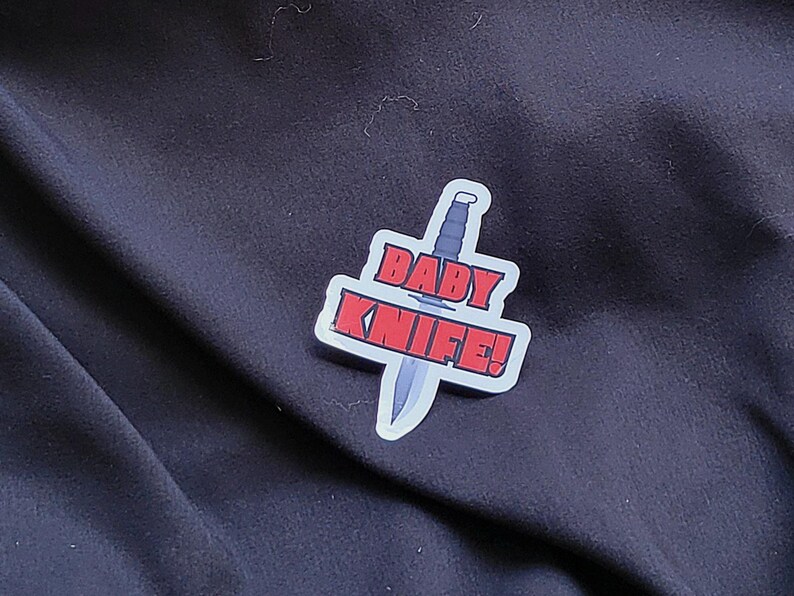 以下が含まれることがあります： 黒い布の背景に、黒いナイフと赤い文字で「BABY KNIFE！」と書かれた白いステッカー