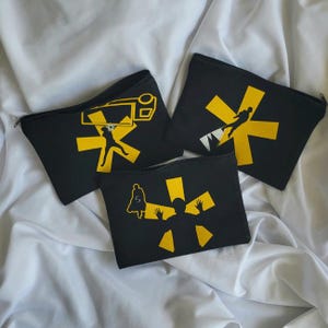 Puede incluir: Tres estuches negros con cremallera y diseños gráficos amarillos. Cada estuche presenta un diseño de silueta diferente, que incluye una persona con los brazos extendidos, una persona con un arma y una persona saltando sobre una montaña. Los estuches están sobre un fondo blanco.