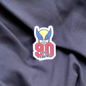 以下が含まれることがあります： 赤と白のステッカーに、数字の90と「til you're」という黒い文字が書かれている。数字の上に、ウルヴァリンの頭部の漫画が描かれている。