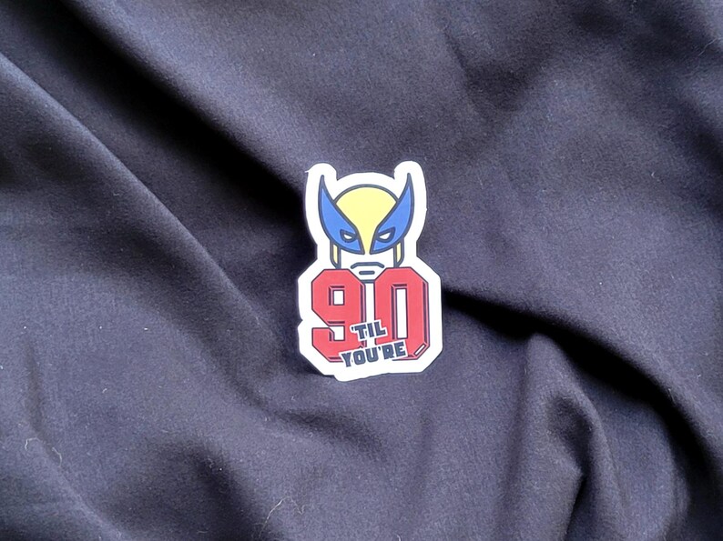 以下が含まれることがあります： 赤と白のステッカーに、数字の90と「til you're」という黒い文字が書かれている。数字の上に、ウルヴァリンの頭部の漫画が描かれている。