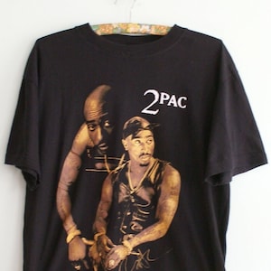 K&ouml;nnte beinhalten: Schwarzes T-Shirt mit einem goldenen Aufdruck von 2Pac. Der Druck zeigt zwei Bilder von 2Pac, eines mit einem schwarzen Bandana und eines ohne. Der Text "2PAC" ist in Wei&szlig; &uuml;ber den Bildern gedruckt.