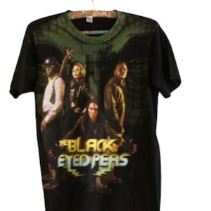 May include: Black t-shirt featuring a group image of The Black Eyed Peas. The band's name is printed in a stylized font with green and yellow accents. The shirt is hanging on a wooden hanger.