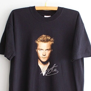 May include: Black t-shirt featuring a portrait of a man with blonde hair and a signature in white. The t-shirt is hanging on a wooden hanger against a white background. The image on the shirt is in shades of brown and gold.