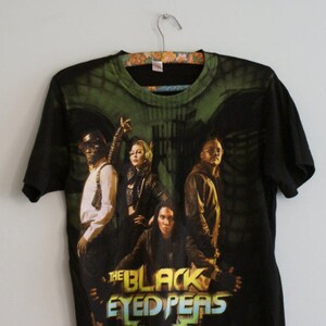 May include: Black t-shirt featuring a graphic of The Black Eyed Peas band members. The design includes the band's name in a colorful, stylized font. The shirt is hanging on a floral patterned hanger.