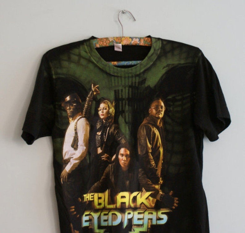 May include: Black t-shirt featuring a graphic of The Black Eyed Peas band members. The design includes the band's name in a colorful, stylized font. The shirt is hanging on a floral patterned hanger.