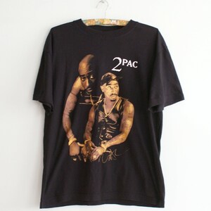 K&ouml;nnte beinhalten: Schwarzes T-Shirt mit einem Grafikdruck von zwei M&auml;nnern, einer mit einem Bandana, beide mit goldenen Ketten. Der Text "2PAC" ist in Wei&szlig; &uuml;ber dem Bild gedruckt.