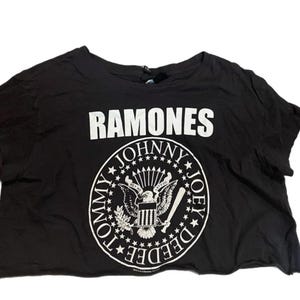 Könnte beinhalten: Schwarzes, kurzes T-Shirt mit dem Wort "RAMONES" in weißen Blockbuchstaben über einem kreisförmigen Grafik. Die Grafik zeigt einen Adler, einen Baseballschläger und die Namen "TOMMY", "JOHNNY" und "DEEDEE".