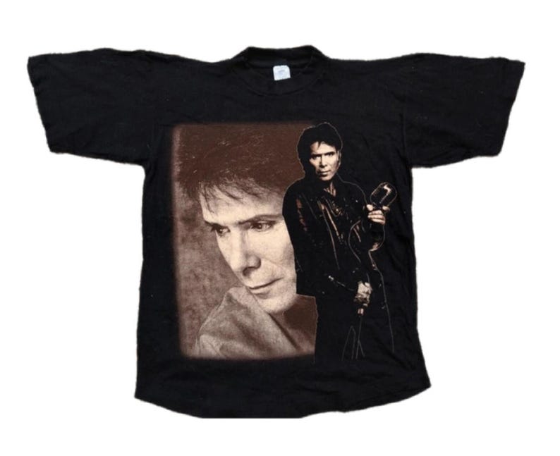 May include: Black t-shirt with a printed image of a man with long dark hair and a microphone. The image is in a brown rectangle.
