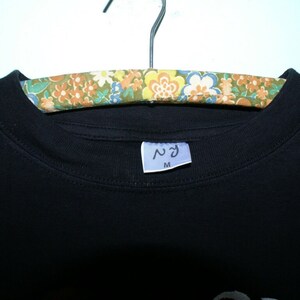 K&ouml;nnte beinhalten: Schwarzes T-Shirt mit einem wei&szlig;en Etikett, auf dem "ヘヴン" und "M" auf einem floral gemusterten Kleiderb&uuml;gel stehen.
