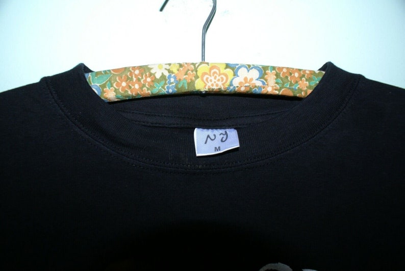 K&ouml;nnte beinhalten: Schwarzes T-Shirt mit einem wei&szlig;en Etikett, auf dem "ヘヴン" und "M" auf einem floral gemusterten Kleiderb&uuml;gel stehen.