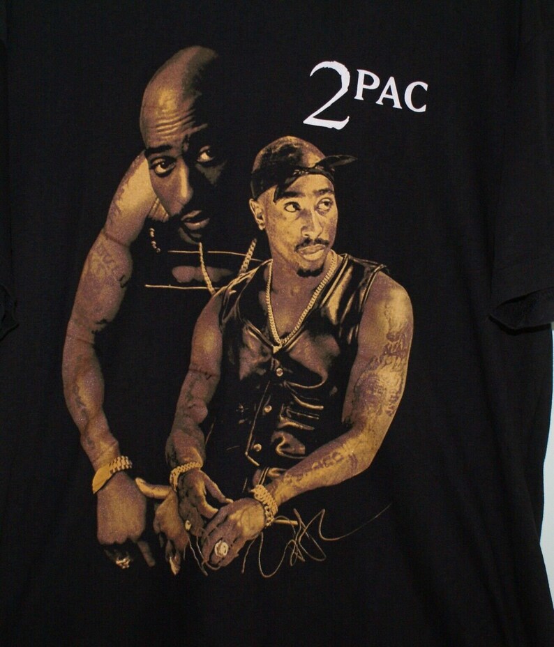 K&ouml;nnte beinhalten: Schwarzes T-Shirt mit einem goldenen Aufdruck von zwei Bildern des Rappers 2Pac. Der Druck enth&auml;lt den Text "2PAC" und eine Unterschrift.