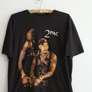 K&ouml;nnte beinhalten: Schwarzes T-Shirt mit einem goldenen Aufdruck von 2Pac. Der Druck zeigt zwei Bilder von 2Pac, eines in einer schwarzen Lederweste und das andere in einem wei&szlig;en Tanktop. Der Text "2PAC" ist in Wei&szlig; &uuml;ber den Bildern gedruckt.